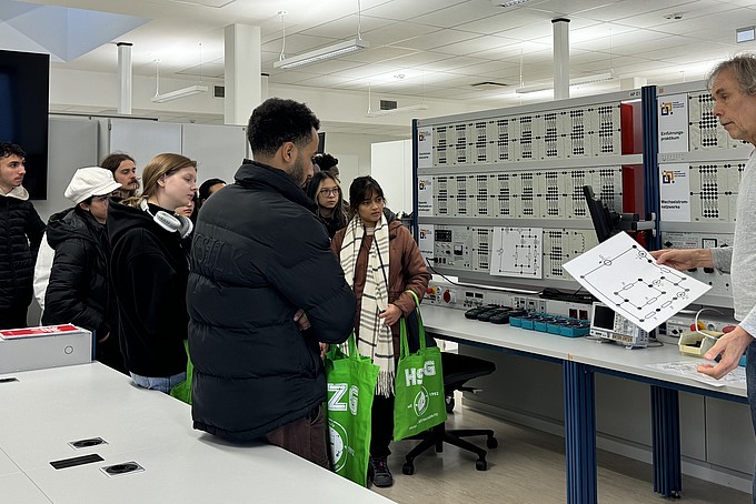 Dr. Wolfgang Menzel explains various aspects of the technical equipment to the visitors in a spacious laboratory of the Faculty of Electrical Engineering and Computer Science' at the HSZG. The laboratory is modernly equipped, with numerous technical boards and devices, while the group listens attentively.