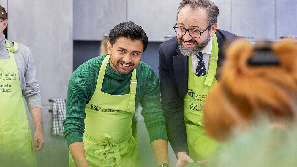 Minister of State and foreign student in aprons cut out cookies with green molds. They lean over the table and smile at the camera.