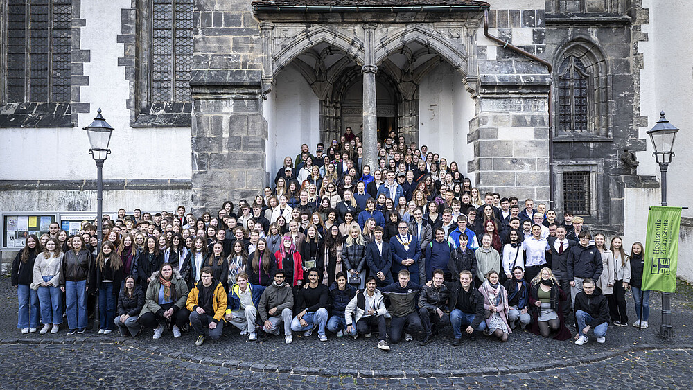 A large crowd of young students and the rector are standing at the side entrance of the church, spread out on the steps and the front square. Everyone is looking into the camera.