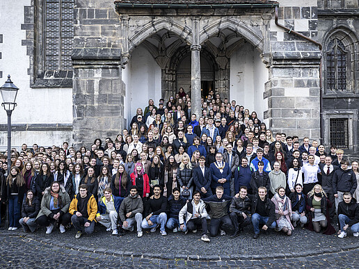 A large crowd of young students and the rector are standing at the side entrance of the church, spread out on the steps and the front square. Everyone is looking into the camera.