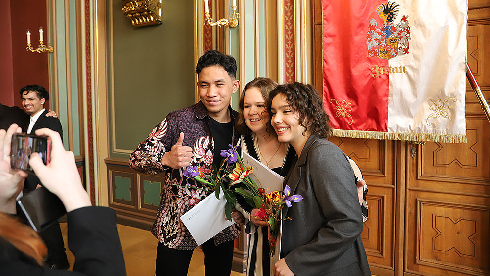 Graduates of the Studienkolleg pose for the camera in front of the Zittau flag in the town hall with their certificates.