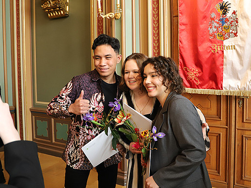 Graduates of the Studienkolleg pose for the camera in front of the Zittau flag in the town hall with their certificates.