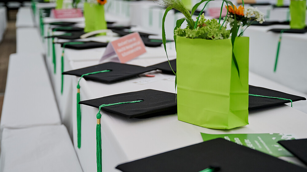 On a table covered with a white tablecloth are graduation hats and bouquets of flowers in green paper bags.