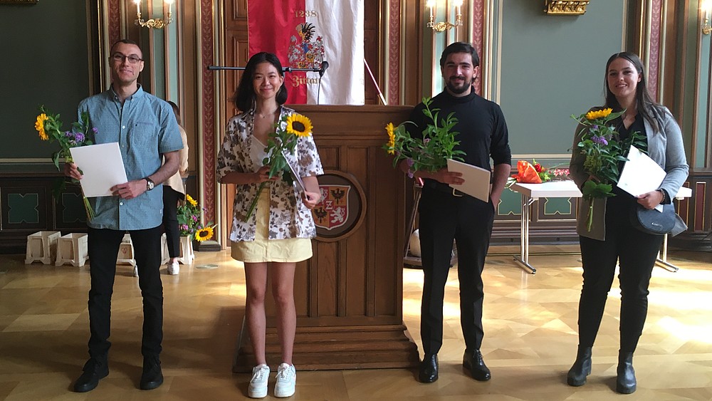 Four Studienkolleg graduates stand in the citizens' hall of Zittau town hall holding their graduation certificates. They smile into the camera as they receive their certificates.